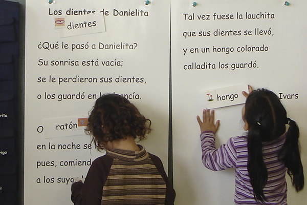 La imagen muestra a dos niñas pegando palabras en un cuento en el pizarrón