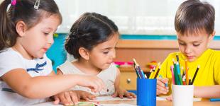 La imagen muestra a dos niñas y un niño realizando una actividad en una mesa escolar