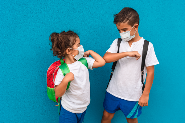 Imagen de dos niños escolares con mascarilla, saludándose con el codo.