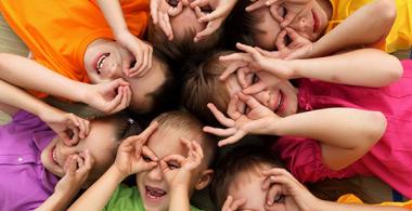 La imagen muestra un grupo de niños acostados en el suelo con  las manos en los ojos